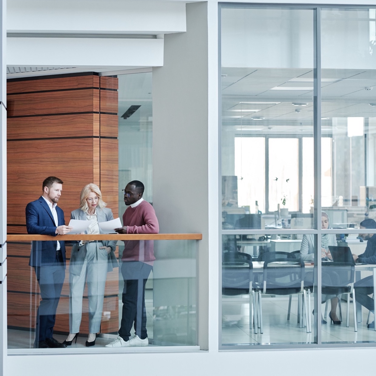 Three colleagues on an office landing, looking over papers — glass offices beyond